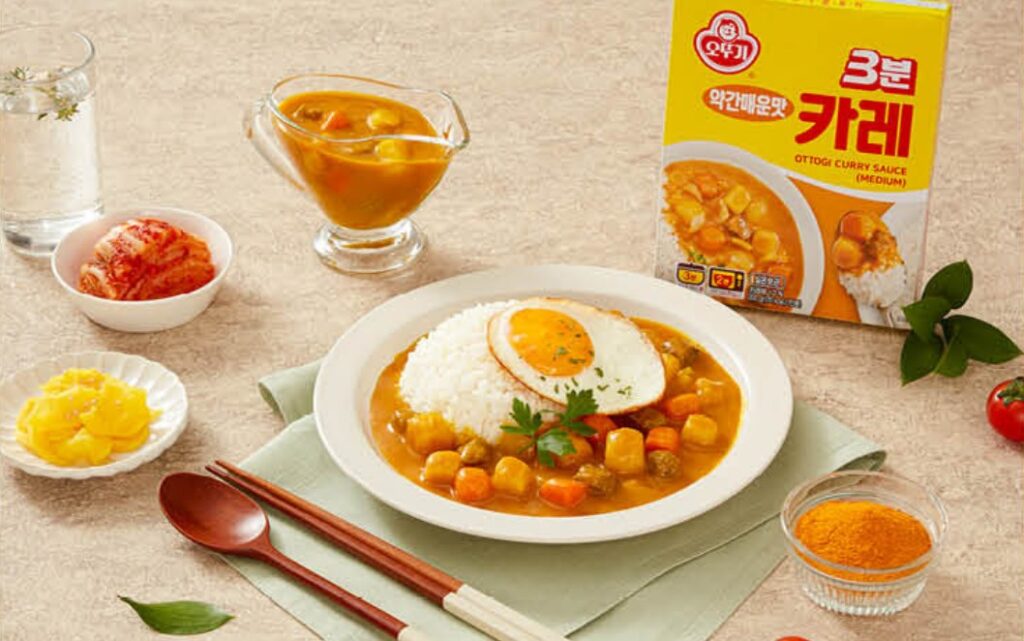 56년 만에 나온 오뚜기 브랜드북, 그 안에 담긴 전략 8 오뚜기 3분 카레로 만든 한 접시 요리 사진. 흰 쌀밥 위에 카레 소스가 넉넉히 뿌려져 있고, 계란 프라이와 파슬리가 고명으로 올라가 있습니다. 주변에는 노란 단무지, 빨간 김치, 카레 가루, 그리고 물 한 잔이 함께 배치되어 있어, 카레를 맛있게 즐기는 한국의 일상적인 식탁 풍경을 연출합니다.