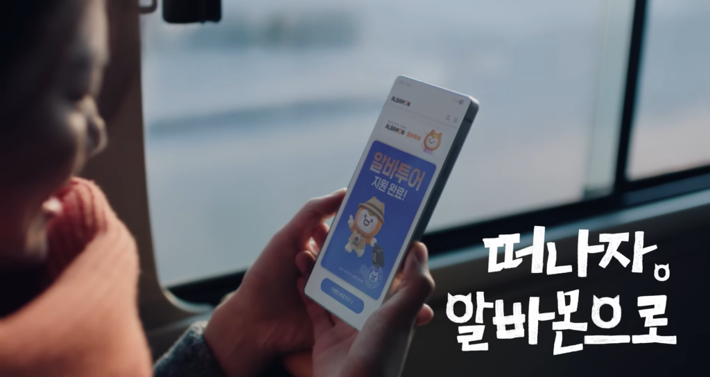 알바몬이 '시급' 대신 '여행'을 제안한 이유 10 알바투어 광고 영상의 한 장면으로, 기차 창가 좌석에 앉아 스마트폰을 보고 있는 모습입니다. 스마트폰 화면에는 알바몬 앱이 실행되어 있고, 탐험가 복장의 모니 캐릭터와 함께 '알바투어 지원 완료!'라는 메시지가 떠 있습니다. 화면 우측 하단에는 '떠나자 알바몬으로'라는 굵은 자막이 배치되어 여행을 떠나는 설렘과 기대감을 표현하고 있습니다.