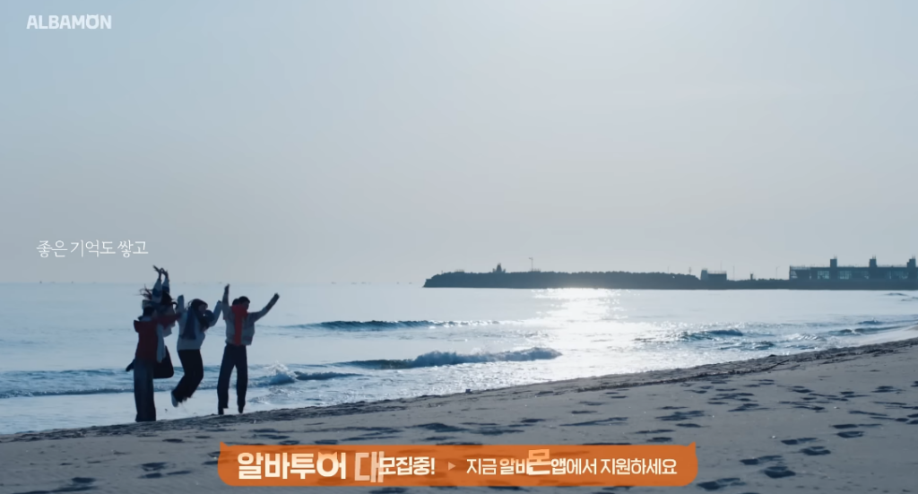 알바몬이 '시급' 대신 '여행'을 제안한 이유 11 탁 트인 부산 바다와 모래사장을 배경으로 네 명의 청년들이 손을 들고 신나게 점프하고 있는 뒷모습입니다. 푸른 하늘과 바다가 시원한 해방감을 주며, 좌측에는 '좋은 기억도 쌓고'라는 자막이, 하단에는 주황색 바에 '알바투어 대모집중! 지금 알바몬 앱에서 지원하세요'라는 안내 문구가 적혀 있습니다.