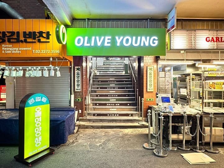 1960년대 올리브영이 있었다면, 아마 이런 모습이었을 거예요 5 광장시장 골목 안에 위치한 올리브영 광장마켓점 입구. 초록색 올리브영 간판 아래 계단에 '올영양행' 문구가 적혀 있으며, 주변 시장 가게들과 나란히 자리하고 있다.